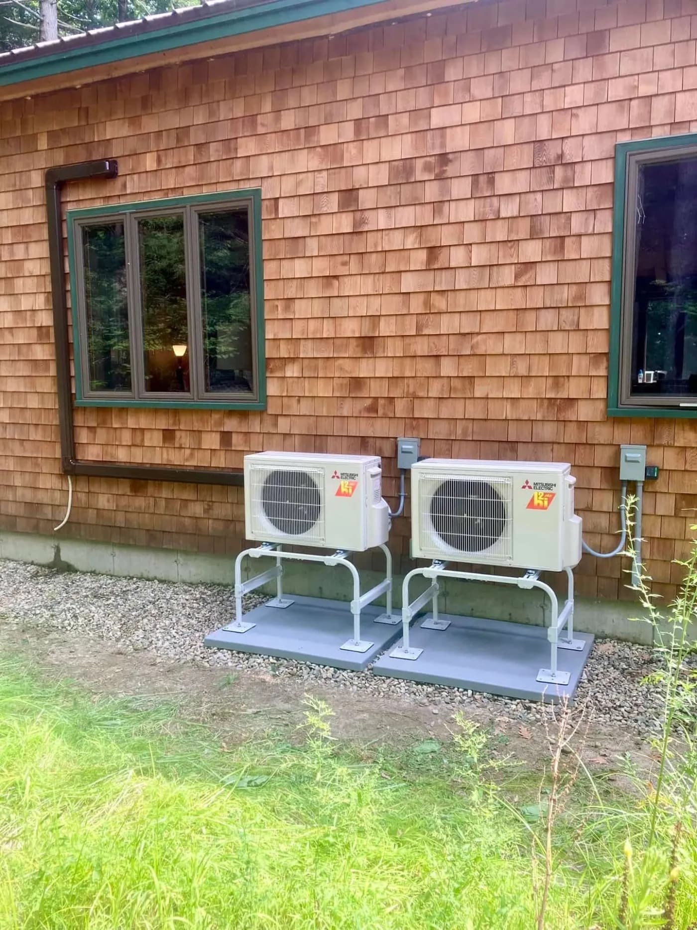 Two Mitsubishi outdoor condensers mounted on elevated stands beside a cedar-shingled Maine home